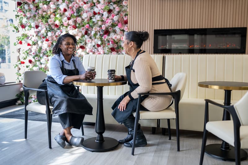 Yefon and a team member in black aprons laughing over coffee at a table by the flower wall