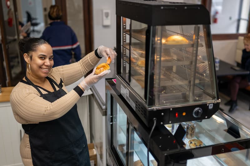 Pulling a warm pastry from the case at the counter, smiling at the camera