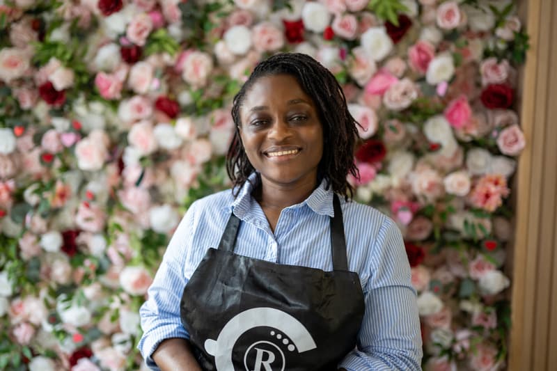 Yefon Fonye, co-owner, smiling in a 96 Royal apron in front of the pink and white rose wall