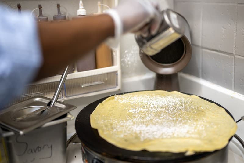 Shaking powdered sugar over a crepe cooking on the iron